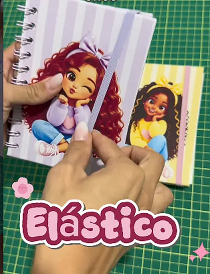 Mãos segurando dois bloquinhos finalizados com elástico, um roxo e um amarelo