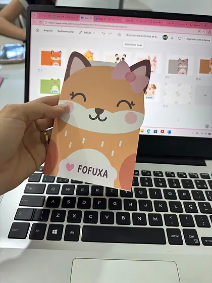 Pessoa segurando um bloquinho impresso com personagem raposa diante da tela do Canva